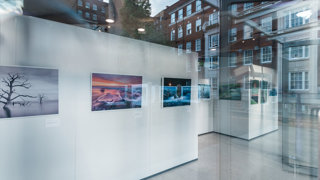 View through a glass window of a photographic exhibition displayed on white panels.