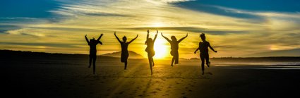 Group of five people jumping on a beach at sunset. The sun is in the front, making the people look like black silhouettes. 