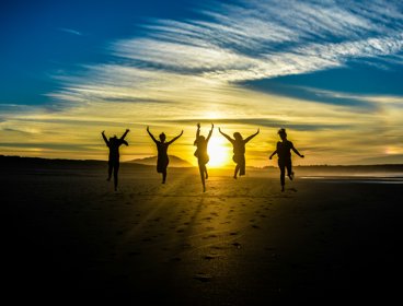 Group of five people jumping on a beach at sunset. The sun is in the front, making the people look like black silhouettes. 