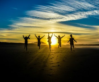 Group of five people jumping on a beach at sunset. The sun is in the front, making the people look like black silhouettes. 