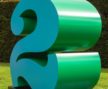 A large green number 2 on a background of grass, with a hedge and cloudy blue skies.