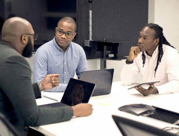 Three people sit at a desk with laptops and a tablet. One sits at the head of the table and the other two opposite each other.