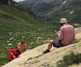A researcher sitting on a rocky slope in a valley writing in a notebook while two others stand testing the rock hardness.