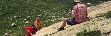 A researcher sitting on a rocky slope in a valley writing in a notebook while two others stand testing the rock hardness.