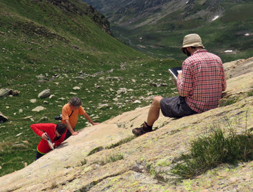 A researcher sitting on a rocky slope in a valley writing in a notebook while two others stand testing the rock hardness.