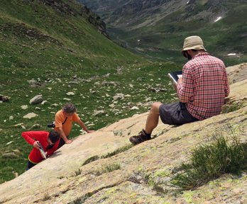 A researcher sitting on a rocky slope in a valley writing in a notebook while two others stand testing the rock hardness.