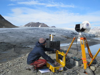 Measurement equipment set up at the base of a glacier. A researcher is crouched down using the equipment.