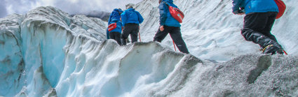 A group of people, all wearing blue jackets and carrying red backpacks, are walking up the side of a glacier. The glacier is tinted blue, and the edges are grey.