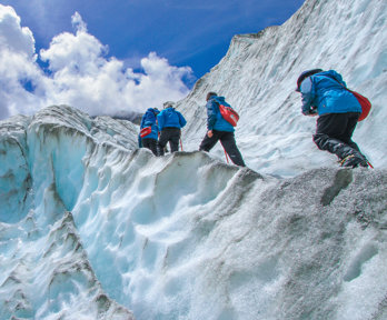 A group of people, all wearing blue jackets and carrying red backpacks, are walking up the side of a glacier. The glacier is tinted blue, and the edges are grey.