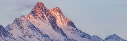 A mountain in the early or late sun, with its tip in soft sunlight. The mountain is purple, blue and white in colour where the rocks and snow meet.
