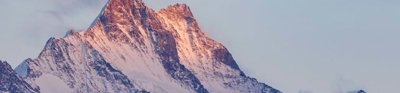 A mountain in the early or late sun, with its tip in soft sunlight. The mountain is purple, blue and white in colour where the rocks and snow meet.