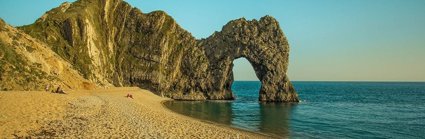 Durdle Door, an arch landscape formation, shows an arch in the rocks at the end of a small coastal sandy bay. The sun is low in the sky casting a soft light onto the rocks and the sea, which is very calm