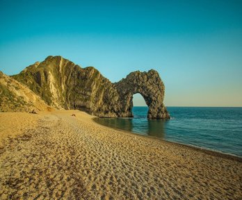Durdle Door, an arch landscape formation, shows an arch in the rocks at the end of a small coastal sandy bay. The sun is low in the sky casting a soft light onto the rocks and the sea, which is very calm