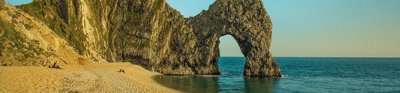 Durdle Door, an arch landscape formation, shows an arch in the rocks at the end of a small coastal sandy bay. The sun is low in the sky casting a soft light onto the rocks and the sea, which is very calm