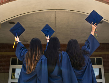 Three students stand before a university campus building in their graduation gowns. They are all holding their graduation caps in the air. 