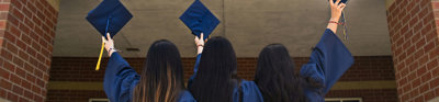 Three students stand before a university campus building in their graduation gowns. They are all holding their graduation caps in the air. 