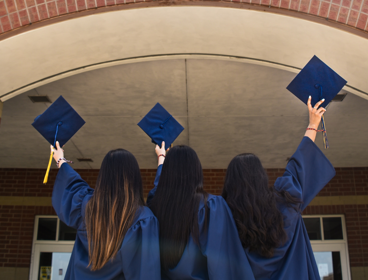Three students stand before a university campus building in their graduation gowns. They are all holding their graduation caps in the air. 