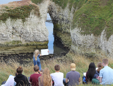 A group of students facing a rocky beach which features an arch landform. The cliffs are chalky and covered in vegetation. There is a teacher facing the group holding up a clipboard