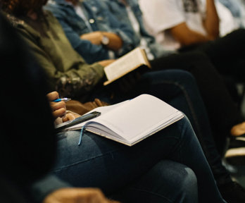 A row of people are seated, some with a notepad and pen on their laps. Others have their arms crossed or resting on their legs.