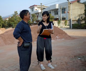 A researcher speaking to a local worker holding a notebook and pen. They are standing in front of a sand heap with a street of houses behind.