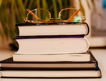 A pair of glasses rest on a stack of five books are stacked on top of each other.