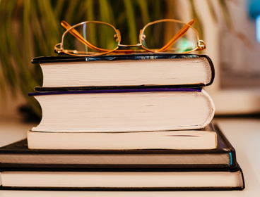 A pair of glasses rest on a stack of five books are stacked on top of each other.