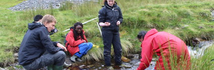 A group of students are gathered by a small stream, one is measuring the depth of a water using a stick. The water is fairly fast moving.