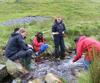 A group of students are gathered by a small stream, one is measuring the depth of a water using a stick. The water is fairly fast moving.