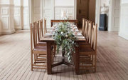 A dark wood dinner table with a long floral centerpiece setup in the middle of the Main Hall.