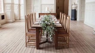 A dark wood dinner table with a long floral centerpiece setup in the middle of the Main Hall.