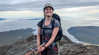 Kathryn Green posing for a photo. She is wearing climbing gear and is standing on the edge of a cliff overlooking a sea.