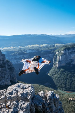 A person base jumping from a tall canyon top, with rocky outcrops and cliffs and trees in the distance