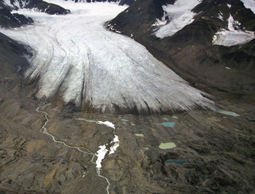 A glacier snout seen emerging from a valley. The glacier is white grey in colour and the valley sides are deep brown and scattered with snow fall