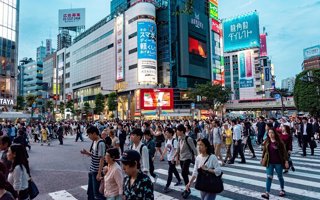 A very busy road crossing in Tokyo, with many people crossing the road at the same time. Zebra crossings can be seen, as well as large buildings with neon lighting in the background