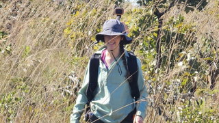 Researcher walks through tall pale brown grass and small trees wearing a rucksack with a LiDAR scanner attached.  