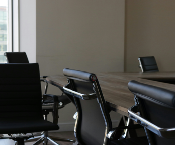 Black conference chairs sit around a square wooden table. One of the chairs sits away from the table unlike the rest