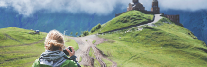 A person stand on a path leading up to a castle on a grassy hill whilst looking into their camera and taking a picture