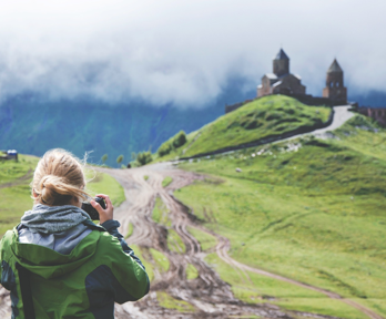 A person stand on a path leading up to a castle on a grassy hill whilst looking into their camera and taking a picture