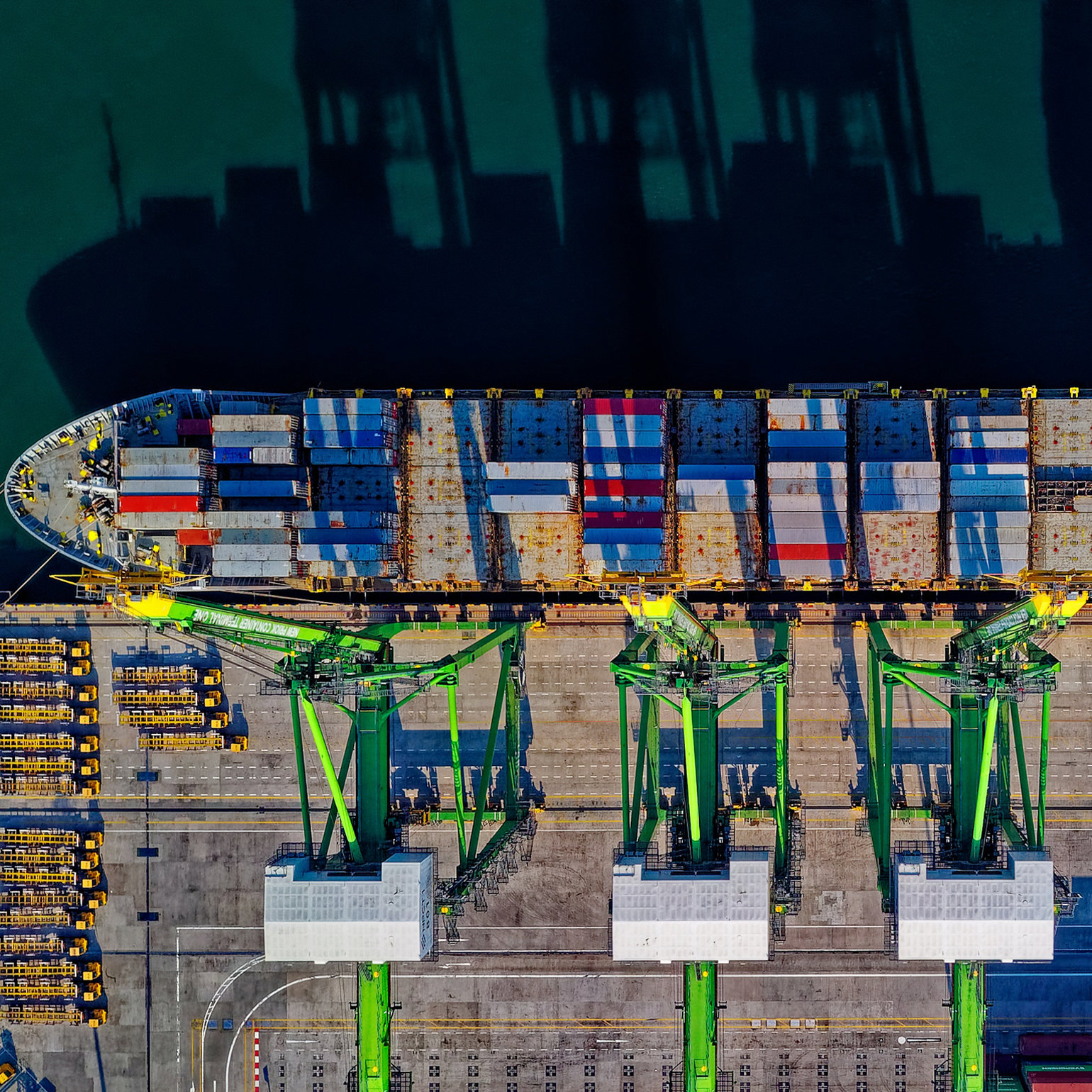 An aerial view of ships at a dockyard with green cranes ready to load them up with containers
