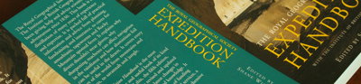 A book titled 'the royal geographical society expidition handbook' lays open but face down on a wooden surface