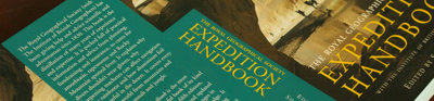 A book titled 'the royal geographical society expedition handbook' lays open but face down on a wooden surface.