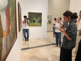 A researcher giving a group of people a tour of an exhibition with paintings on the walls. A photographer is stood in the corner taking photos of the group.