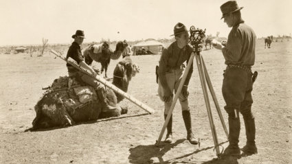 Sepia photo of a crew mapping borders in the desert. One person, who's looking directly at the camera, sits on a camel and holds a large measuring stick.