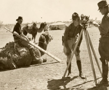 Sepia photo of a crew mapping borders in the desert. One person, who's looking directly at the camera, sits on a camel and holds a large measuring stick.