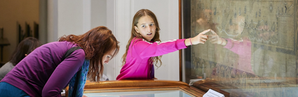 A person hunched over a display cabinet and a young child pointing at an old world map.