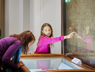 A person hunched over a display cabinet and a young child pointing at an old world map.