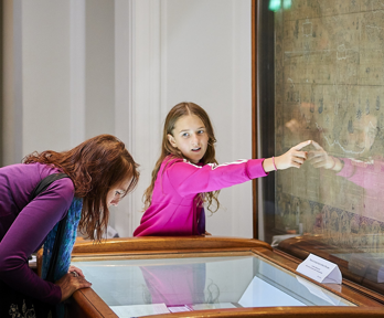 A person hunched over a display cabinet and a young child pointing at an old world map.