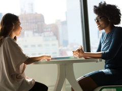 Two people sit across from each other at a small white table. They appear to be mid conversation with their hands resting on the table. 