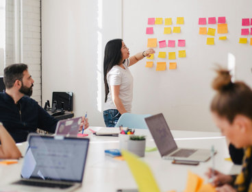 People sat around a table typing on laptops. One person is stood at the front pointing at square sticky notes stuck on a whiteboard. 