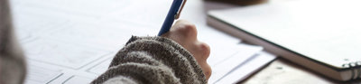 Person sat at a wooden table writing on paper near a white ceramic mug.